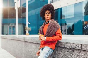 Young curly mixed-race boy drinks coffee on the street against the background of office buildings. Youth culture and fashion. Student photo