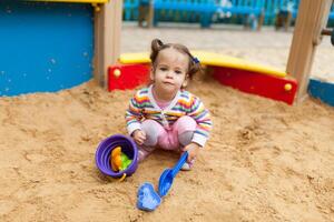 a little girl with two tails is dressed in a striped colorful jacket is playing in the sandbox on the playground photo