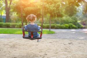 Unrecognizable Little Caucasian girl riding swing at playground sunny summer day rear view photo