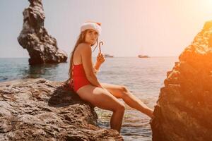 Woman travel sea. Happy tourist enjoy taking picture on the beach for memories. Woman traveler in Santa hat looks at camera on the sea bay, sharing travel adventure journey photo