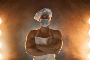 nuevo normal concepto. muscular cocinero vistiendo protector médico mascarilla, posando con doblada brazos en ahumado antecedentes foto