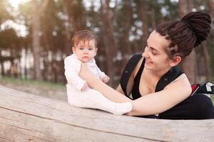 A beautiful smiling young brunette woman with long dreadlock hair hold a pretty baby. Mother looks tenderly at the child. They are in the green summer forest. photo