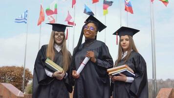 Three young women university graduates of different nationalities smile and stand together holding books and a diploma in their hands against many flags of different countries video