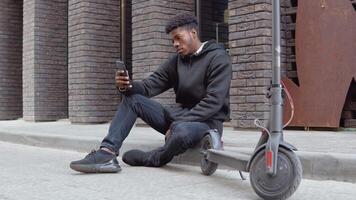 A young African American man in a black sweater and black jeans and sneakers sits on the sidewalk near a building with a dark brick facade. Urban lifestyle of young people video