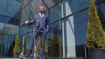 un africain américain dans une classique bleu affaires costume des stands avec un électrique scooter sur le rue près une moderne miroir bâtiment avec le sien main dans le sien poche. bas vue dans plein longueur video