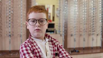 A red-haired boy in a plaid red and white shirt stands with transparent glasses for vision correction turned his shoulder to the camera with and folds his hands in the lock video