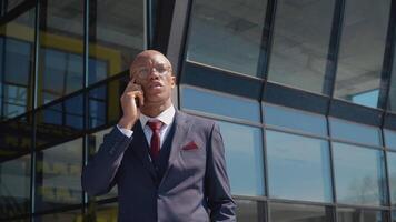 Young attractive african american businessman talking on the phone near a modern mirror building. Holding smartphone, surfing networks. Modern technology and communication video