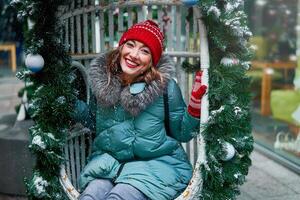 Young beautiful caucasian girl dressed in a warm winter jacket, knitted hat and gloves sits on Christmas decorated swing outdoor photo
