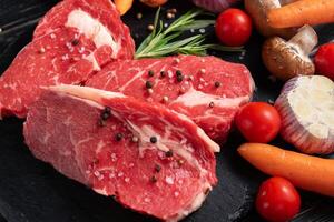 Three pieces of juicy raw beef with vegetables on a stone cutting board on a black wooden table background. photo