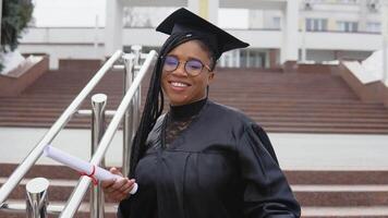 A young woman at the university at the master's mantle holds a diploma and corrects her glasses while looking at the camera. Portrait in front of the central entrance to the university on background video