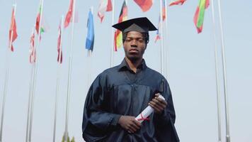 jong Afrikaanse Amerikaans mannetje afstuderen staand in voorkant van de camera met een diploma en boeken in haar handen. de leerling staat buiten met de Internationale vlaggen Aan achtergrond. video