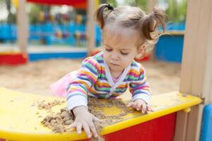 a little girl with two tails is dressed in a striped colorful jacket is playing in the sandbox on the playground photo