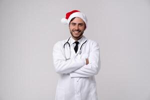 Young handsome doctor in white uniforme and Santa Claus hat standing in studio on white background loking at camera abd teeth smiling photo