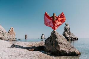 Woman travel sea. Young Happy woman in a long red dress posing on a beach near the sea on background of volcanic rocks, like in Iceland, sharing travel adventure journey photo