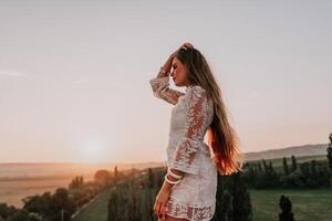Happy woman in white boho dress on sunset in mountains. Romantic woman with long hair standing with her back on the sunset in nature in summer with open hands. Silhouette. Nature. Sunset. photo
