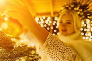 A blonde woman in white dress and a crown of gold ornaments decorate Christmas tree with gold ornaments and lights. The tree is decorated with gold balls and is lit up with lights. photo