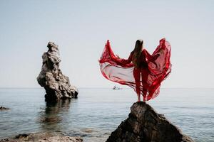 Woman travel sea. Young Happy woman in a long red dress posing on a beach near the sea on background of volcanic rocks, like in Iceland, sharing travel adventure journey photo