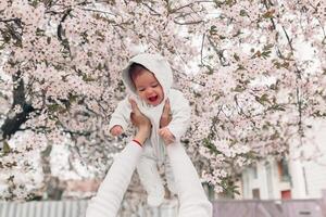 Portrait of happy joyful child in white clothes over tree flowers blossom background. Family playing together outside. Mom cheerfully hold little daughter photo