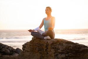 Woman practice yoga lotus pose to meditation with summer vacation rocky beach happiness and relaxation. Calm female exercise with yoga meditate ocean beach with sunset golden time. photo