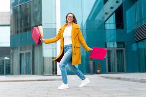 Business Woman With Shopping Bags Dressed Yellow Coat Jumping With Happiness Outdoors Corporative Building Background photo