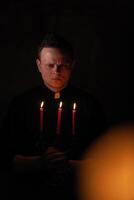 retrato de hermoso católico sacerdote o pastor con perro cuello, oscuro rojo antecedentes. con Tres rojo vela en el mano foto