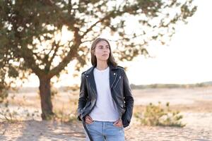 Young beautiful caucasian woman dressed in a leather jacket and jeans stands on the beach on a wood background. photo