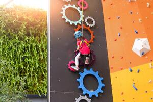 niño alpinismo en pared en diversión centro. alpinismo formación para niños. pequeño niña en vestido alpinismo engranaje escalada alto. extremo activo ocio para niños. foto