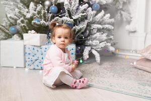 hermosa linda pequeño niña sentado en el piso cerca el nuevo año árbol. fiesta ánimo, infantil espontaneidad y dulce atmósfera foto