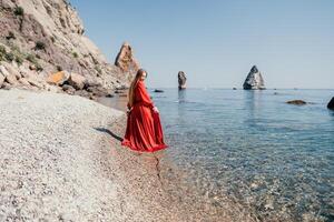 Woman travel sea. Happy tourist in red dress enjoy taking picture outdoors for memories. Woman traveler posing on the rock at sea bay surrounded by volcanic mountains, sharing travel adventure journey photo