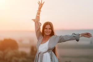 Happy woman standing with her back on the sunset in nature in summer with open hands. Romantic beautiful bride in white boho dress posing with mountains on sunset photo