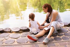 pequeño caucásico niña sentado siguiente a su madre en el apuntalar de un artificial lago en el parque. madre con hija en un caminar en un soleado verano día foto