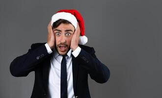 Young handsome caucasian guy in business suit and Santa hats stands on gray background in studio very surprised hands on cheeks. photo
