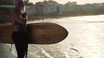 un vieux homme avec une barbe et une planche de surf des stands sur le plage video