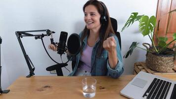 une femme Faire Podcast portant écouteurs et une casque est séance à une bureau avec une ordinateur écran video