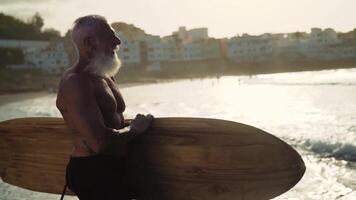 een oud Mens met een baard en een surfboard staat Aan de strand video
