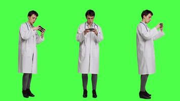 General practitioner playing video games on mobile phone app, having fun with online gaming competition against greenscreen backdrop. Cheerful doctor relaxing with internet game. Camera A.