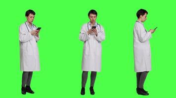 Physician navigating on social media apps to text people, stands against greenscreen backdrop. Medic in white coat browsing websites online, making checkup appointments. Camera A. video