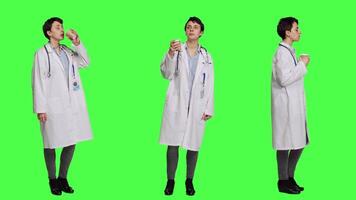 Happy medic serving hot coffee cup as refreshment against greenscreen backdrop, wearing white coat and drinking caffeine beverage. General practitioner enjoying drink, medical expertise. Camera A. video