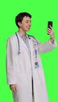 Side view Medic specialist having remote videocall conversation with patients on smartphone internet connection, greenscreen backdrop. Doctor discusses about disease with people on videoconference. Camera B. video