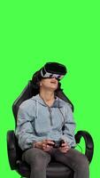 Front view Woman playing video games on console with controller and vr headset, sitting against greenscreen backdrop. Gamer having fun with online tournament, virtual reality interactive cyberspace. Camera B.