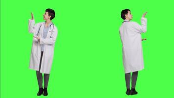 Physician pointing to the left or right to create web commercial standing against greenscreen backdrop, doing a marketing advertisement in studio. Woman medic in white coat shows an ad. Camera A. video