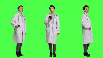 Woman specialist looking at phone watch and waiting for patients to arrive at exam appointments, greenscreen backdrop in studio. General practitioner in white coat acting impatient. Camera A. video