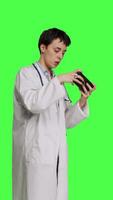 Side view General practitioner playing video games on mobile phone app, having fun with online gaming competition against greenscreen backdrop. Cheerful doctor relaxing with internet game. Camera B.