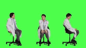 Physician in white coat waiting for patients at consultations, feeling impatient sitting on a chair against greenscreen backdrop. Medic practitioner with stethoscope waits for people. Camera A. video