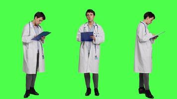 Physician taking notes after patient examination against greenscreen backdrop, writing down medicaments to cure illness. Doctor in white coat using clipboard papers for checkup. Camera A. video