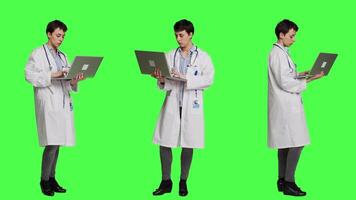 Confident physician searching for new medicaments to treat disease, using laptop to browse on online web pages against greenscreen backdrop. Medic in white coat checks internet sites. Camera A. video