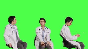 Physician in white coat waiting for patients at consultations, feeling impatient sitting on a chair against greenscreen backdrop. Medic practitioner with stethoscope waits for people. Camera B. video