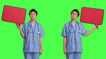 Medical assistant holding a red speech bubble icon against greenscreen, creating advertisement with empty copyspace billboard sign. Nurse shows cardboard poster, marketing announcement. Camera B. video