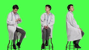 General practitioner acting impatient sitting on a chair against greenscreen backdrop, waiting for patients to attend checkup appointments. Woman specialist in white coat waits for people. Camera A. video