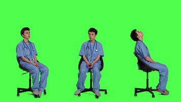 Medical assistant is waiting for patients to attend exam appointments, feeling impatient while she sits in a chair. Nurse with stethoscope and scrubs waits for people against greenscreen. Camera A. video